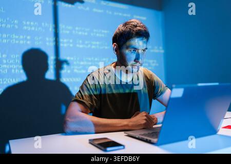 Focused programmer concentrating on laptop, debugging code with projected lines on wall background, developing new software, working late in dark tech Stock Photo