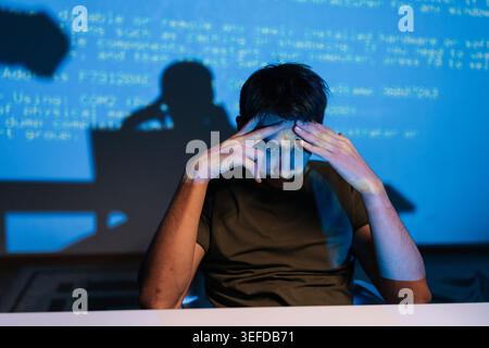 Stressed young male programmer facing challenging coding problem, feeling frustrated while working on laptop with lines of blue code projecting on Stock Photo