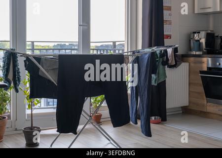 Laundry dries on a folding metal clothes rack indoors next to a balcony window in an apartment kitchen. Stock Photo