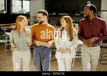 Four individuals stand together in an office space, engaged in a conversation. They hold tablets and appear to share ideas. The setting has large wind Stock Photo