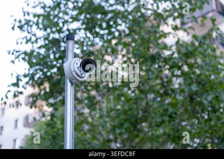 White surveillance camera is mounted on a metal pole in front of a green tree in a residential neighborhood. Stock Photo
