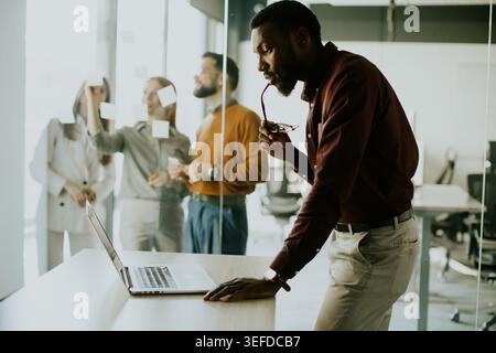 A man stands by a desk, looking at a laptop while holding his glasses. In the background, a team collaborates, discussing ideas with sticky notes on a Stock Photo