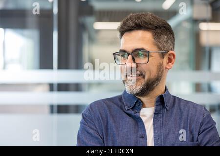 Confident and successful business man wearing glasses and a denim shirt, smiling and looking away, showcasing professionalism and a positive attitude in a corporate office setting Stock Photo