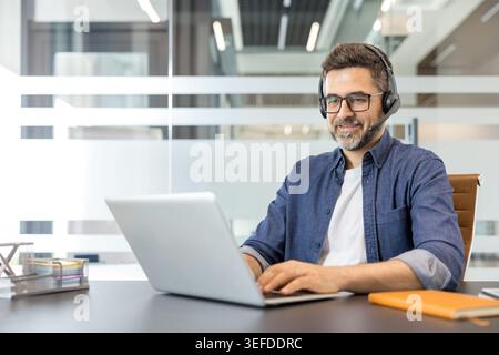 Mature man working on a laptop in a modern office, wearing a headset and smiling, representing remote work, online communication and a positive work environment Stock Photo