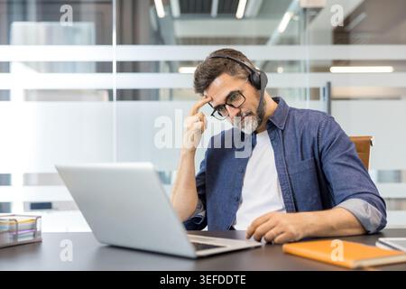 Frustrated middle-aged man feeling tired and experiencing work stress, touching his forehead, while working remotely on a laptop in a modern office setup Stock Photo