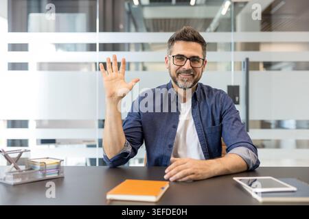 Mature businessman with glasses smiling and waving hand during a video conference, communicating and greeting online from his modern office workplace environment Stock Photo