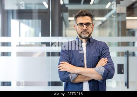 Confident mid adult businessman standing with arms crossed, wearing glasses and a denim shirt in a modern office, representing professionalism and leadership Stock Photo