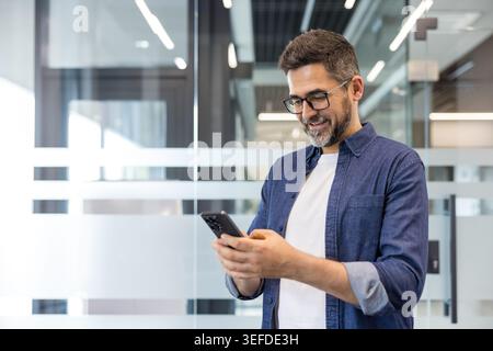 Smiling man with glasses using smartphone in a modern office, networking and texting during the workday, confident professional connecting via mobile apps and social media, waist-up portrait Stock Photo