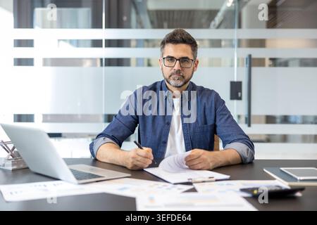 Businessman in a modern office writing notes and reviewing financial reports, representing professionalism, strategy, finance, and successful business operations Stock Photo