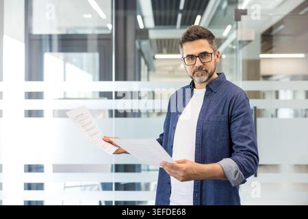 Confident businessman wearing denim collared shirt and eyeglasses standing in a contemporary office corridor, holding and reviewing important business documents Stock Photo