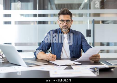 Professional businessman in glasses sits at a modern office desk, focused on reading and signing documents with a pen beside a laptop and financial charts, serious and productive Stock Photo