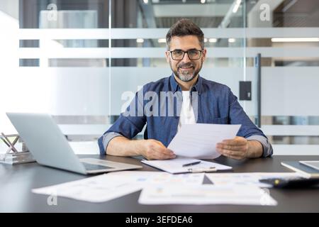 Confident businessman in glasses and denim shirt smiles at camera while seated at desk with laptop and financial reports, holding documents in a modern office setting Stock Photo