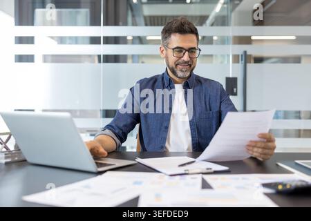Professional man wearing glasses and a denim shirt sitting at his office desk, happily engaged in work while using a laptop and reviewing financial documents with graphs on the table Stock Photo