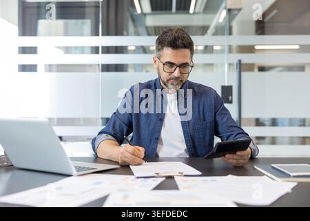 Mature professional man wearing eyeglasses working on finances, holding a calculator, writing on paper, and reviewing documents at an office desk with a laptop and scattered papers Stock Photo
