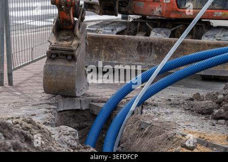 Close up of excavator bucket working on underground utility installation with blue protective conduit pipes inside an urban construction trench. Detai Stock Photo