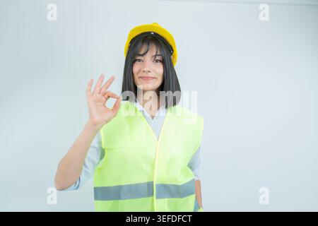 Portrait of young female engineer or construction worker making OK gesture while wearing yellow safety helmet and reflective vest. Studio shot symboli Stock Photo
