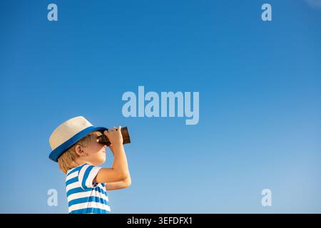 Child looking through binoculars against blue sky. Kid having fun outdoor in summer. Adventure and travel concept Stock Photo