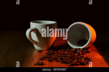 Coffee beans spilling from an overturned white espresso cup, with another cup standing nearby on a warm illuminated wooden table, creating a dramatic Stock Photo