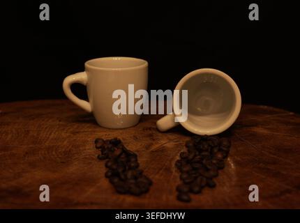 Two white ceramic espresso cups sit on a textured wooden surface next to scattered dark roasted coffee beans, creating a rustic and natural coffee sce Stock Photo