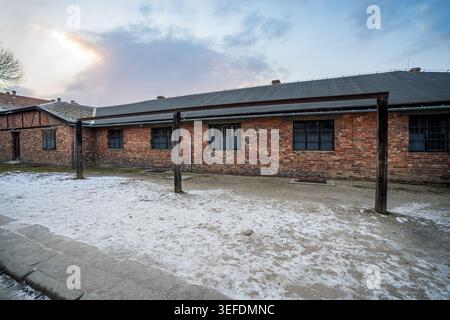 Linear facade of a red brick barracks building at Auschwitz I. In front of the structure stand three tall vertical wooden posts joined by a horizontal Stock Photo