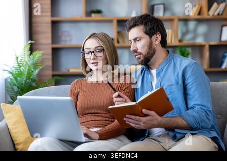 Young couple working together on a laptop, discussing plans, writing notes in a notebook, and sharing ideas while sitting comfortably on a sofa in their home office Stock Photo