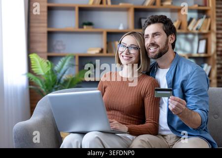 Young happy couple making an online purchase, sitting on a couch at home, using a laptop and holding a credit card for e-commerce transactions and digital banking Stock Photo