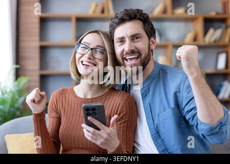 Happy couple at home celebrating great news on a smartphone, smiling and fist-pumping in the living room after a joyful online notification of success and achievement Stock Photo