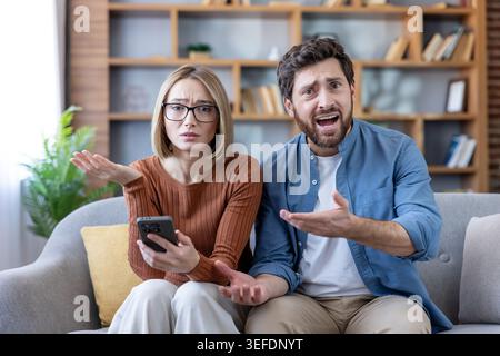 Shocked young couple sitting on a sofa at home, feeling frustrated and confused while looking at a smartphone, reacting with anger and miscommunication to unexpected information Stock Photo