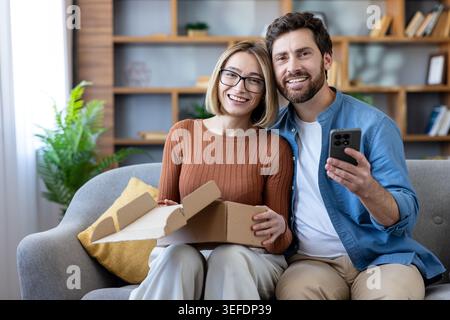 Happy young couple smiling and sitting on a sofa in their living room, opening a cardboard box with a new purchase while using a smartphone, symbolizing successful e-commerce and home delivery Stock Photo
