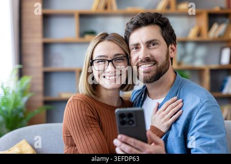 Happy couple smiling and looking at the camera while taking a selfie together on a modern smartphone, embracing each other in their cozy living room at home Stock Photo