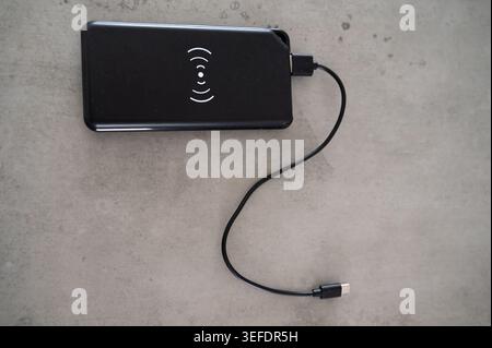 Black power bank with cable resting on concrete surface, wireless charging icon visible, braided usbc lead coiled, textured finish, minimalist tech ac Stock Photo