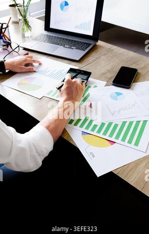 Person analyzes colorful charts on a wooden desk. Their hands move between papers and a smartphone. Laptop displays a pie chart nearby. Focus on data Stock Photo