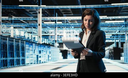 Data center manager verifying infrastructure status with clipboard files and holographic visuals. Server room worker using audit charts and blue wireframe simulation to oversee equipment Stock Photo