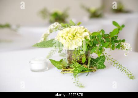 Festive flower decoration in white and green on bar tables with hydrangea blossom, twigs, herbs and ivy in a glass jar with moss, copy space, selected Stock Photo