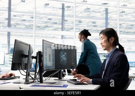 Senior hardware developer in semiconductor manufacturing research facility designing microchip layout using PC. Elderly Asian man working at office desk, optimizing PCB network topology using computer Stock Photo