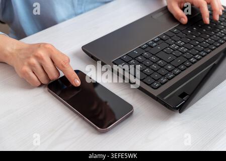 Closeup of smartphone and laptop on office desk with woman interacting on touchscreen, representing mobile communication and digital workflow Stock Photo