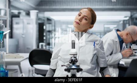 Fatigued biologist stretches in medical lab after using scientific microscope for cellular analysis. Tired scientist relaxes back at workbench, examines sample with optical device gear, camera B Stock Photo