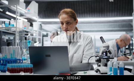 Sick lab expert rubbing head temples, feeling ache while working on laptop, studying genomics. Fatigued laboratory technician hurting while studying DNA strand mapping on notebook, camera A Stock Photo