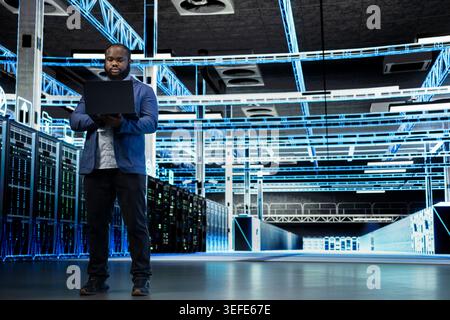 Data center engineer uses laptop to manage IT infrastructure rigs used for database storage. African american man in server farm monitoring internet cloud computing equipment rigs, technology concept Stock Photo
