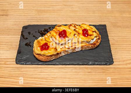 Open toast with goat cheese rolls, red jam and balsamic drizzle arranged on black slate over light wood. Stock Photo