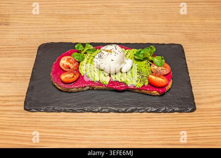 Open toast topped with pink beetroot spread, sliced avocado, burrata, cherry tomatoes and greens on black slate over wood. Stock Photo