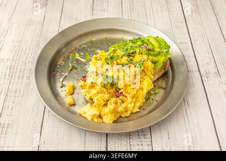 Creamy scrambled eggs and sliced avocado on toast topped with chives and microgreens sits on a silver plate over wood.** Stock Photo