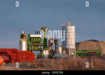 An asphalt plant with silos, machinery, and construction materials under clear sky in industrial setting Stock Photo