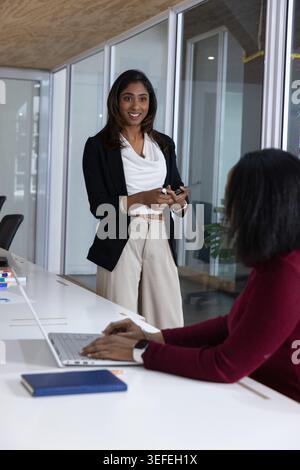 Diverse women in businesswear using tablet stylus while peer typing laptop near blue planner office Stock Photo