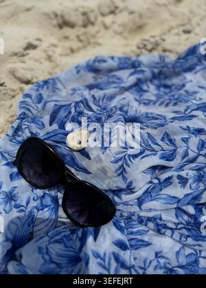 Black designer sunglasses placed on a blue and white floral patterned silk sarong scarf on the beach sand. Stock Photo