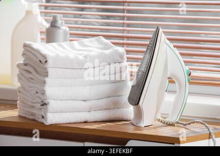 Modern iron with stack of clean towels on commode near window in bathroom Stock Photo