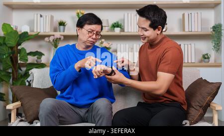 Asian son helping senior father learn to use smartwatch in living room. Stock Photo