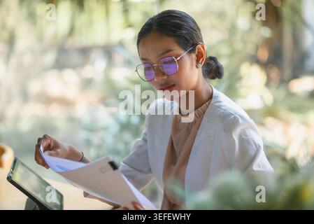 Businesswoman in glasses reviews printed reports beside a tablet at a wooden table, representing financial research, document analysis, data planning, Stock Photo