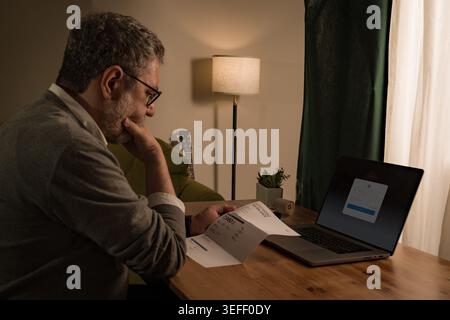 Senior man sitting at home at night reviewing bills and expenses, looking worried while checking financial documents and planning payments on laptop w Stock Photo