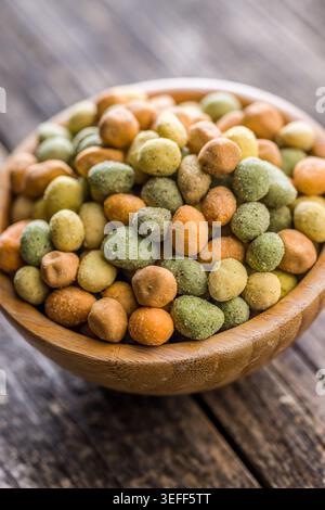 Rustic wooden bowl heaped with mixed coated peanuts and crunchy wasabi peas on a weathered wooden table, evoking casual snacking, texture, and savory Stock Photo